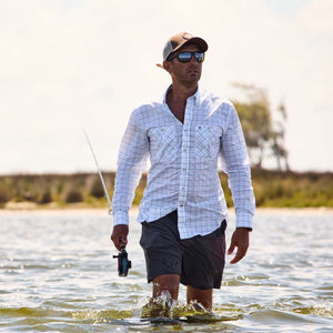 A man in Tom Beckbes Crenshaw Performance Shorts wades through waist-deep water, fishing under a partly cloudy sky.