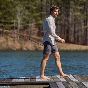 A man in Crenshaw Performance Shorts by Tom Beckbe strolls barefoot on a dock by the lake, holding a fishing rod.