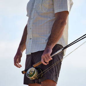 Man in Crenshaw Performance Short and shirt by Tom Beckbe, holding a fishing rod against a pale sky backdrop.