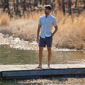 Man on a wooden dock by a lake, wearing a checkered shirt and Crenshaw Performance Short from Tom Beckbe.