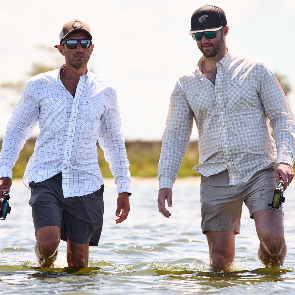 Two men in Crenshaw Performance Shorts and polyester hats wade through water with fishing rods under a clear sky.