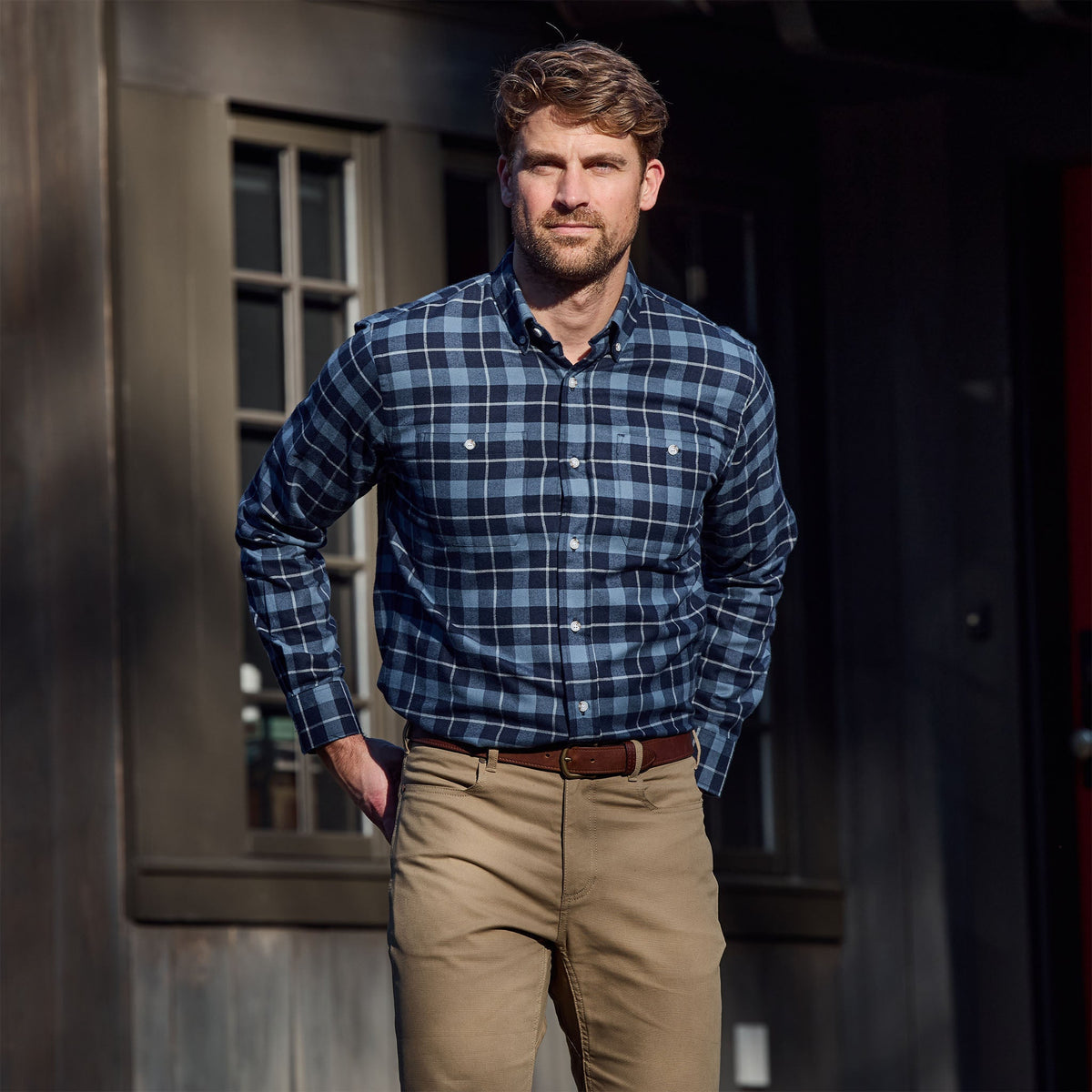 Man wears the Tom Beckbe Dixon Brushed Cotton Twill Shirt and khaki pants outside a dark wooden building with windows.