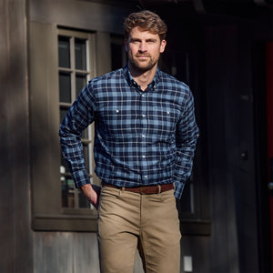 Man wears the Tom Beckbe Dixon Brushed Cotton Twill Shirt and khaki pants outside a dark wooden building with windows.
