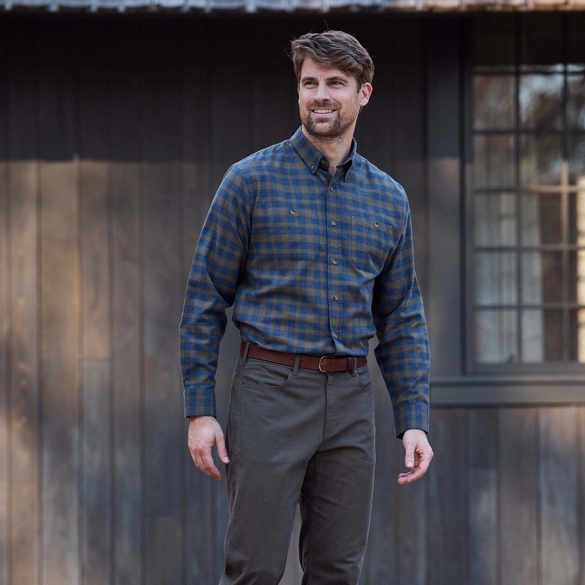 Man wears Tom Beckbe Dixon Brushed Cotton Twill Shirt with olive pants outside a dark wooden building.