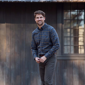 Man in a Tom Beckbe Dixon Brushed Cotton Twill Shirt and dark pants smiles outdoors by a wooden building.