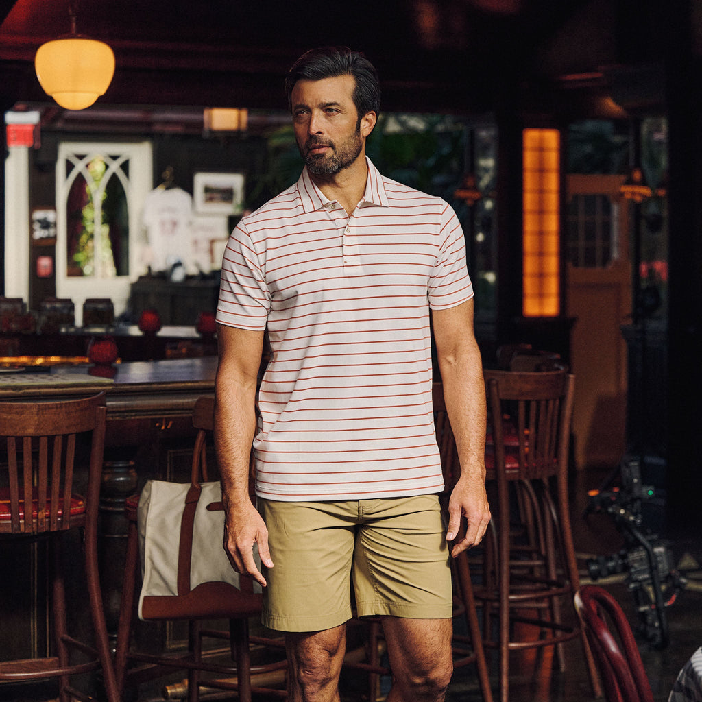 A man in a Tom Beckbe Hutton Performance Polo and khaki shorts stands in a dimly lit bar with wooden chairs.