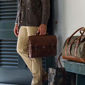 A person in khaki pants holds a Tom Beckbe Leather Messenger Bag with a padded laptop sleeve by a bench.