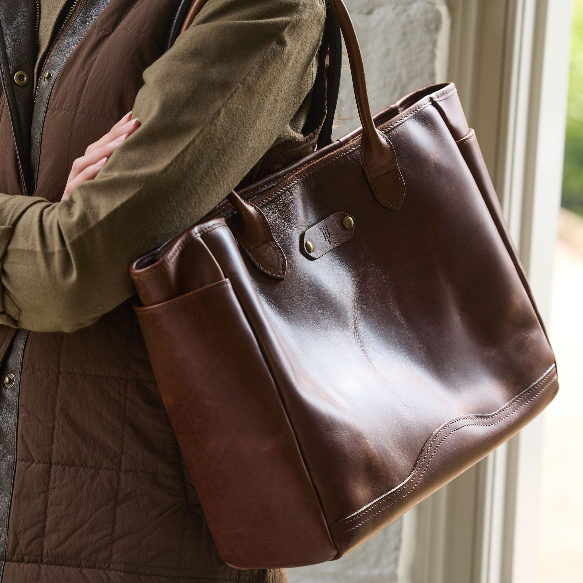 A person carries the Tom Beckbe Palmer Leather Tote, a durable bag with two handles and metal hardware.