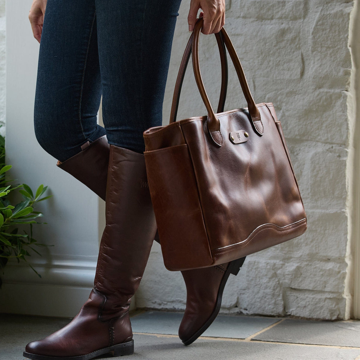 Person holding the Tom Beckbe Palmer Leather Tote, wearing jeans and brown knee-high boots by a white wall.