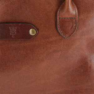 Close-up of Tom Beckbes Palmer Leather Tote, featuring embossed logo, strap, and brass button detail on the front.