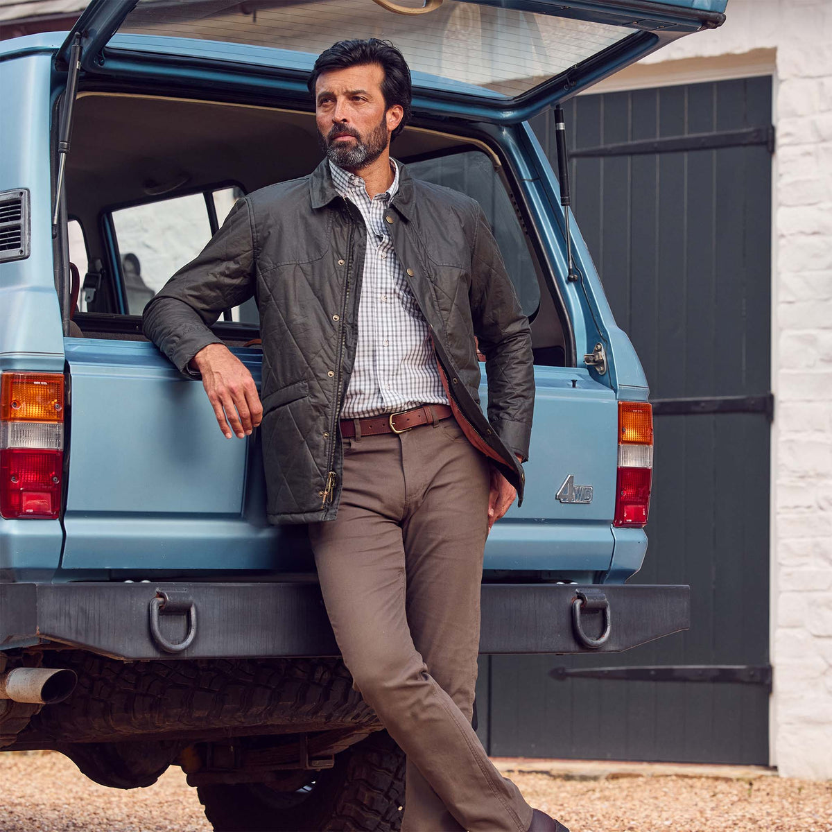 A man in a Tom Beckbe Paddock Quilted Jacket leans on the trunk of a blue SUV outside a white building with black doors.