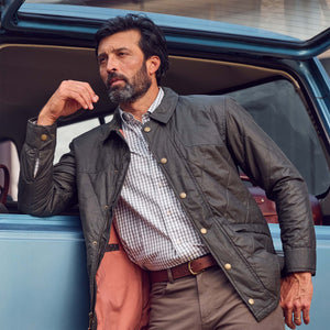 A man in a checkered shirt and Tom Beckbe Paddock Quilted Jacket leans against an open car door, hand near his face.