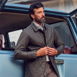 A man in a Tom Beckbe Paddock Quilted Jacket adjusts his zipper beside an open blue SUV.