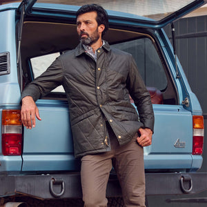 A man in a Tom Beckbe Paddock Quilted Jacket and tan pants leans on a blue SUV with its tailgate open.