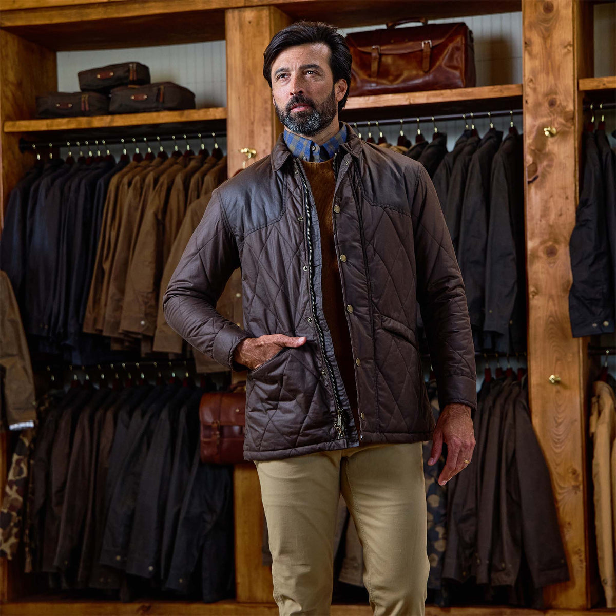 A man in a Tom Beckbe Paddock Quilted Jacket and beige pants stands in a store with weatherproof jackets behind him.
