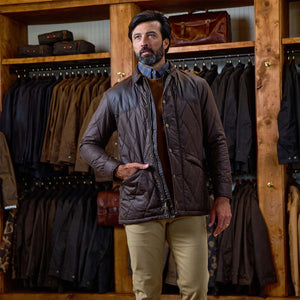 A man in a Tom Beckbe Paddock Quilted Jacket and beige pants stands in a store with weatherproof jackets behind him.