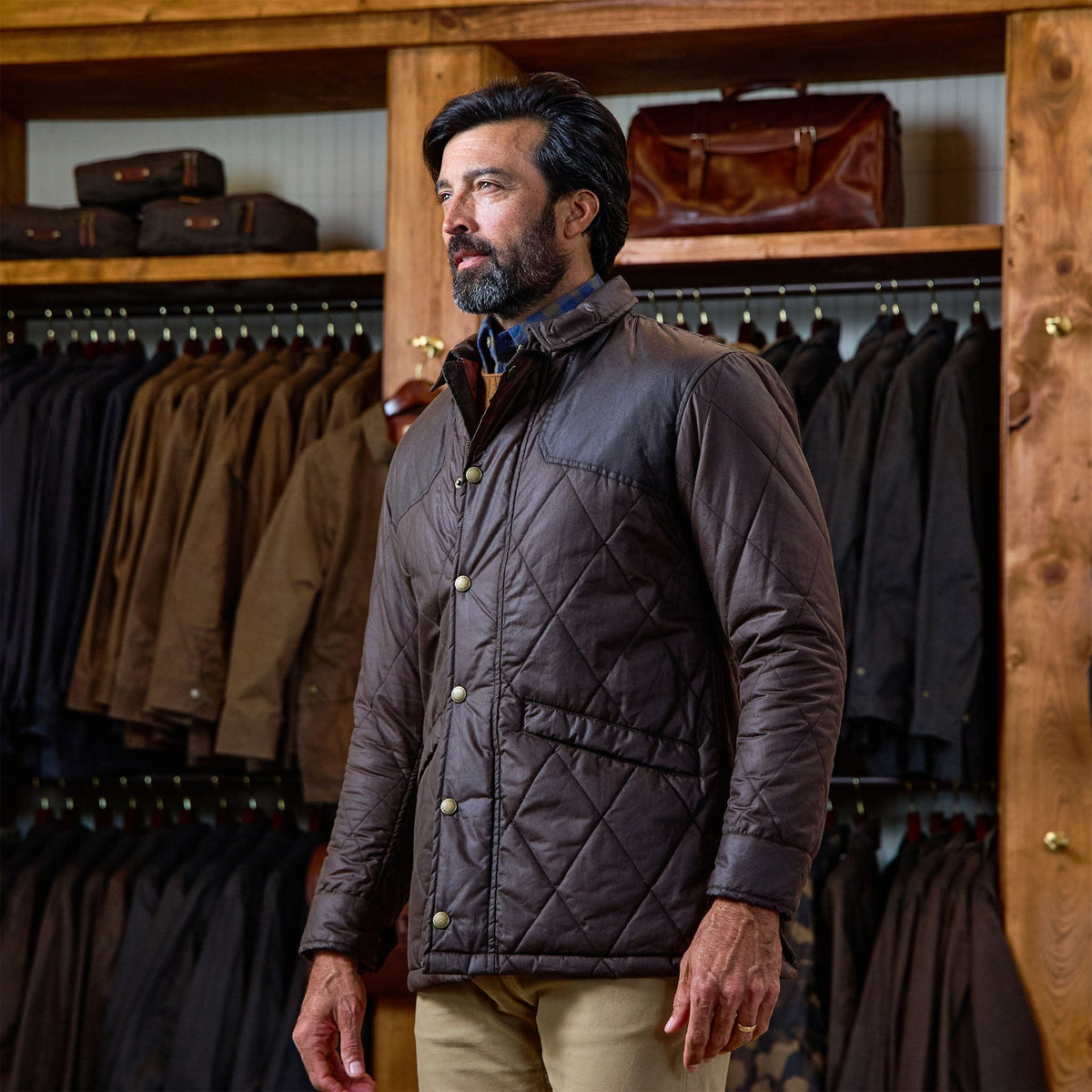 Man wears a Tom Beckbe Paddock Quilted Jacket in a store with weatherproof jackets and bags in the background.