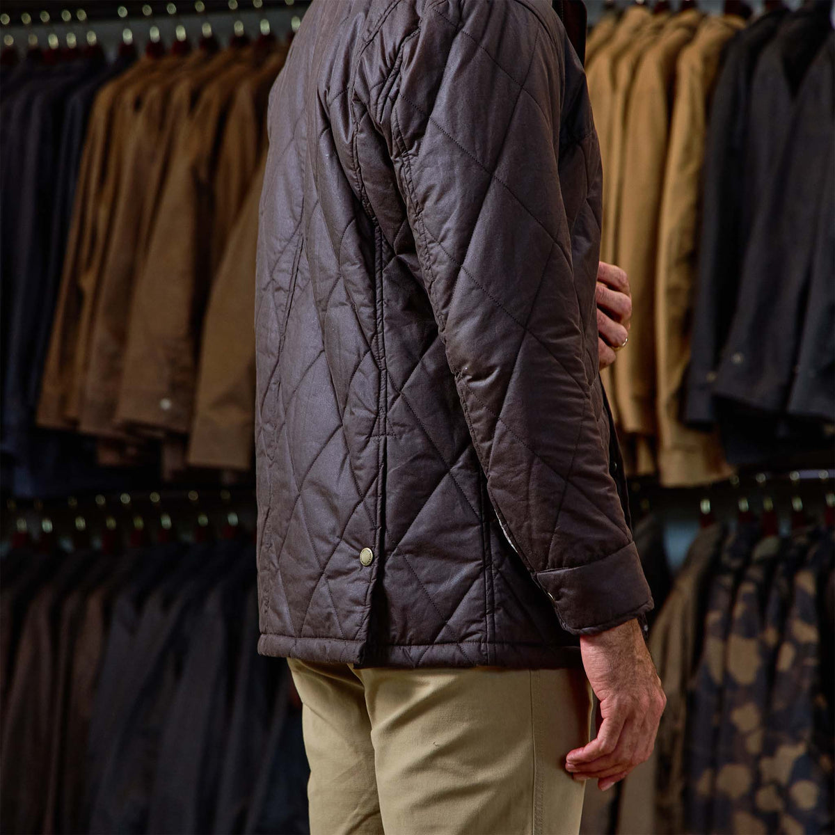 Someone in a Tom Beckbe Paddock Quilted Jacket and beige pants stands before racks of weatherproof, black jackets.