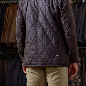 A person wears the Tom Beckbe Paddock Quilted Jacket with beige pants, standing by a clothing rack.
