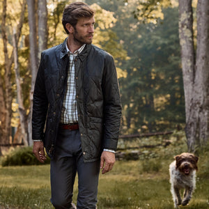 A man in a Tom Beckbe Paddock Quilted Jacket strolls through the woods with his dog trotting behind.