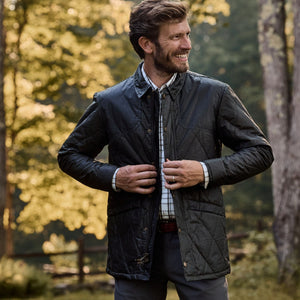 Man in a Tom Beckbe Paddock Quilted Jacket stands outdoors, smiling and adjusting his breathable jacket among trees.