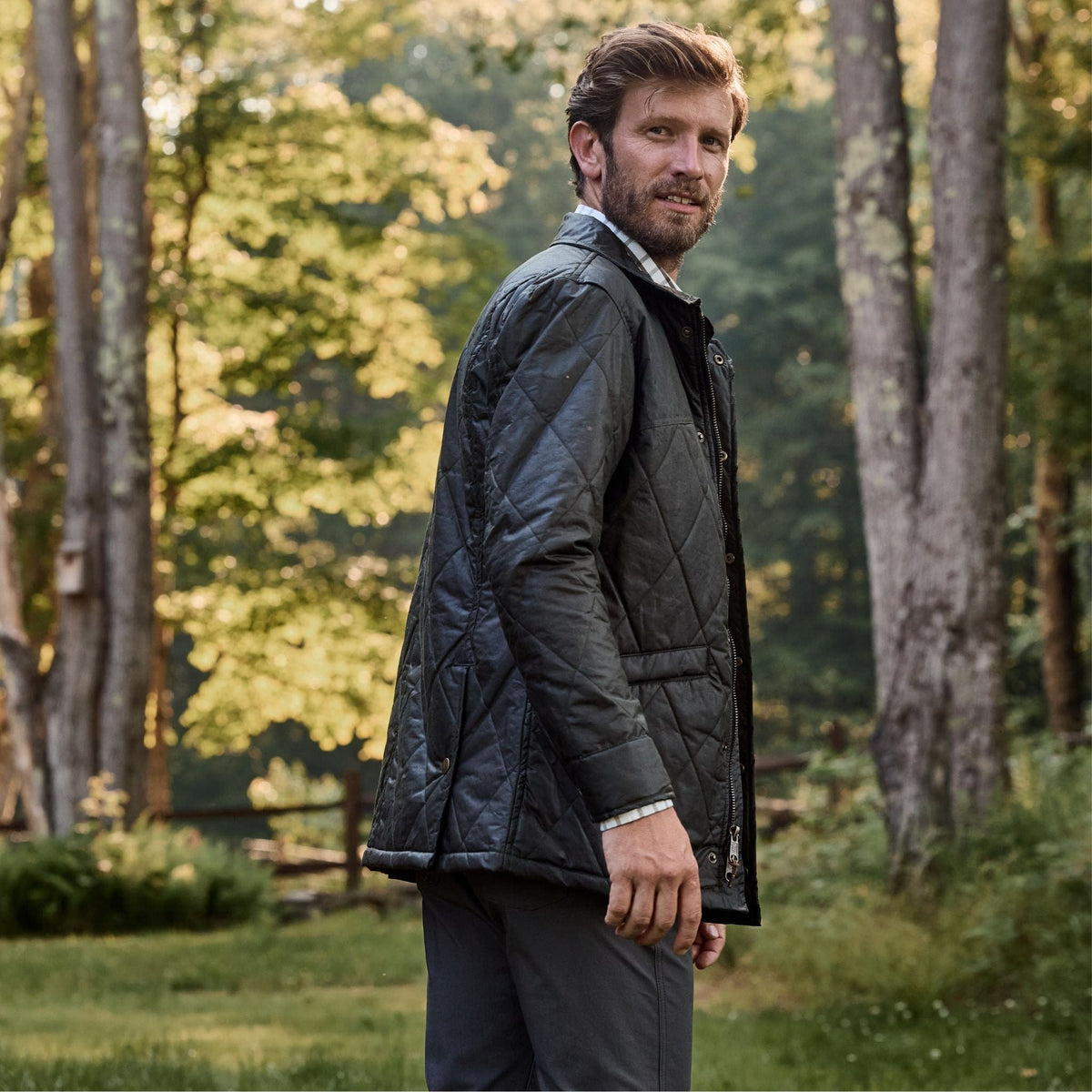 A man models the Tom Beckbe Paddock Quilted Jacket in a forest setting surrounded by trees and greenery.