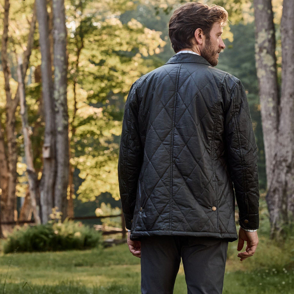 Man in a Tom Beckbe Paddock Quilted Jacket with breathable insulation stands in forest, sunlight filtering through leaves.
