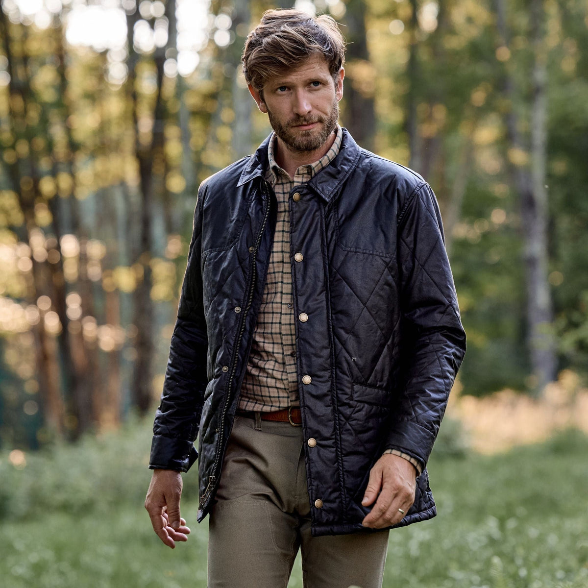 A man in a Tom Beckbe Paddock Quilted Jacket walks through a grassy area with trees in the background.