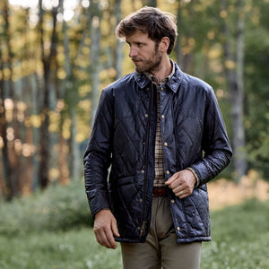A man wearing a Tom Beckbe Paddock Quilted Jacket stands in a forest, showcasing rugged outdoor wear and gazing left.