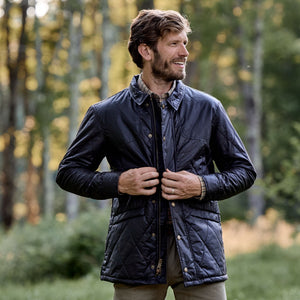 A man buttons a Paddock Quilted Jacket by Tom Beckbe outdoors with trees in the background.
