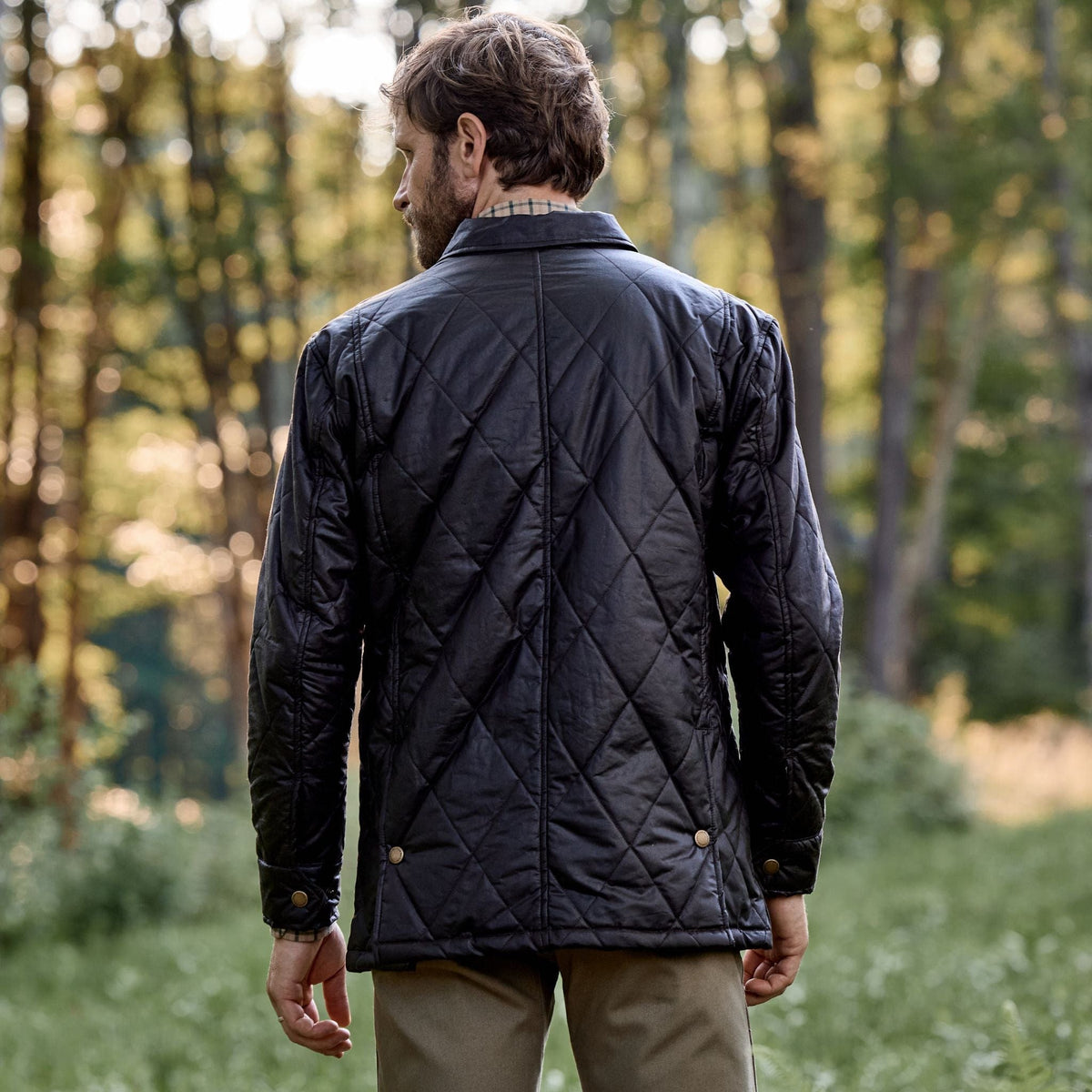 A man in a Tom Beckbe Paddock Quilted Jacket stands amidst the forest greenery, facing away from the camera.