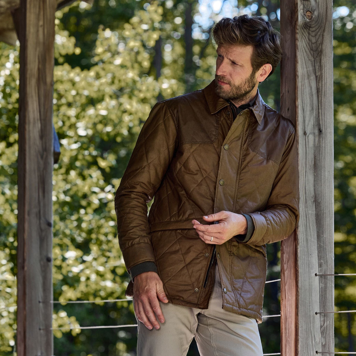A man in a Tom Beckbe Paddock Quilted Jacket leans against a post outdoors, with trees in the background.