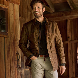 A man in a Tom Beckbe Paddock Quilted Jacket and beige pants smiles by a wooden cabin door.