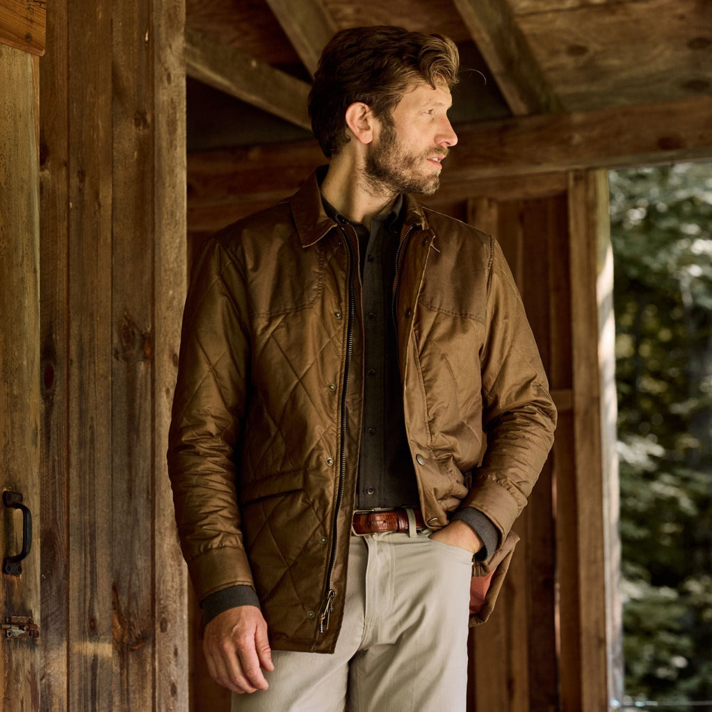 A man in a Tom Beckbe Paddock Quilted Jacket and light pants stands on a porch, glancing sideways with trees behind.