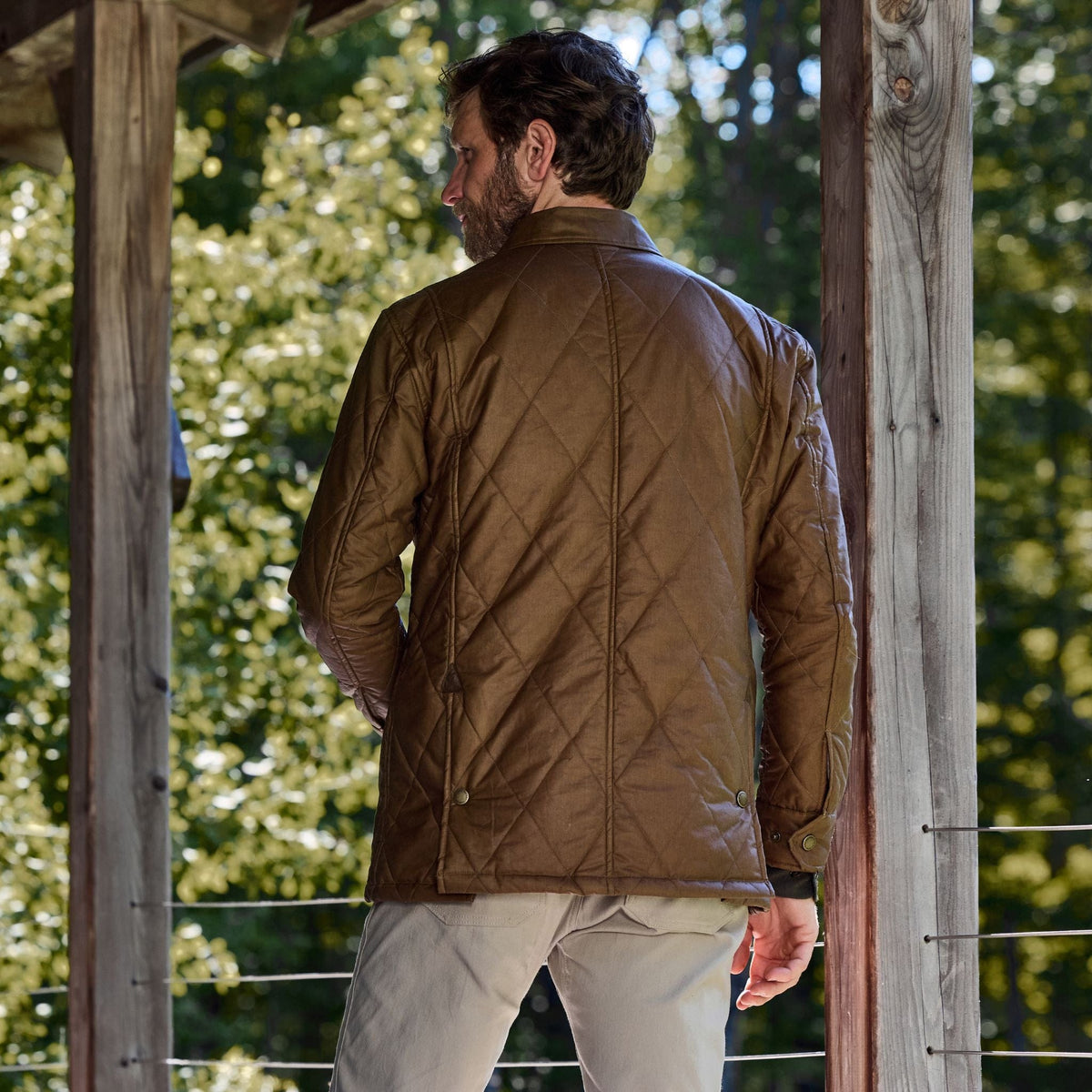 Man on a porch in Tom Beckbes Paddock Quilted Jacket and beige pants, trees in the background.