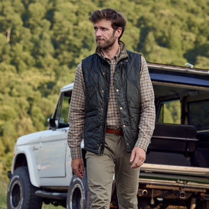 A man in a plaid shirt and Tom Beckbes Paddock Quilted Vest stands by a white SUV amid scenic trees.
