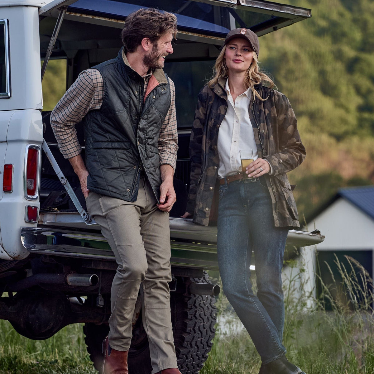 Two people in outdoor gear, featuring a Tom Beckbe Paddock Quilted Vest, lean against an SUV with trees and a house behind.
