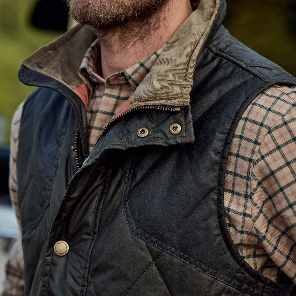 A person in a Tom Beckbe Paddock Quilted Vest over a plaid shirt stands outdoors.