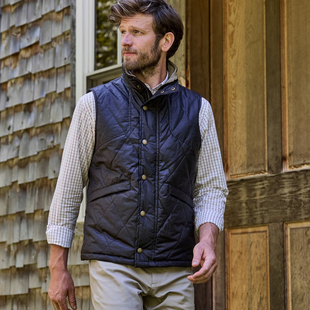 A man in a Tom Beckbe Paddock Quilted Vest and plaid shirt stands outside a wooden building, looking left.