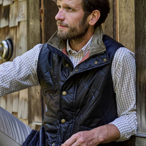 A reflective man in a checkered shirt and Tom Beckbes Paddock Quilted Vest sits outdoors, with a wooden building silhouette.
