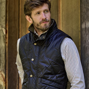Man in a Tom Beckbe Paddock Quilted Vest and plaid shirt leans against a wooden wall, gazing to the side.