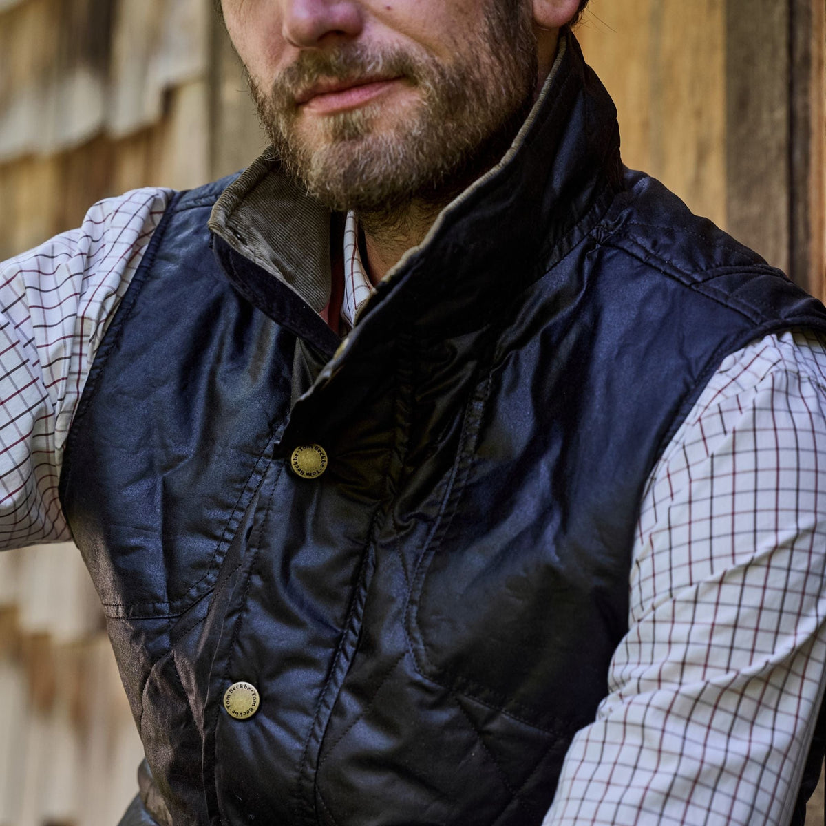 Man in a Tom Beckbe Paddock Quilted Vest and checkered shirt stands outdoors by wooden wall, prepped for any weather.
