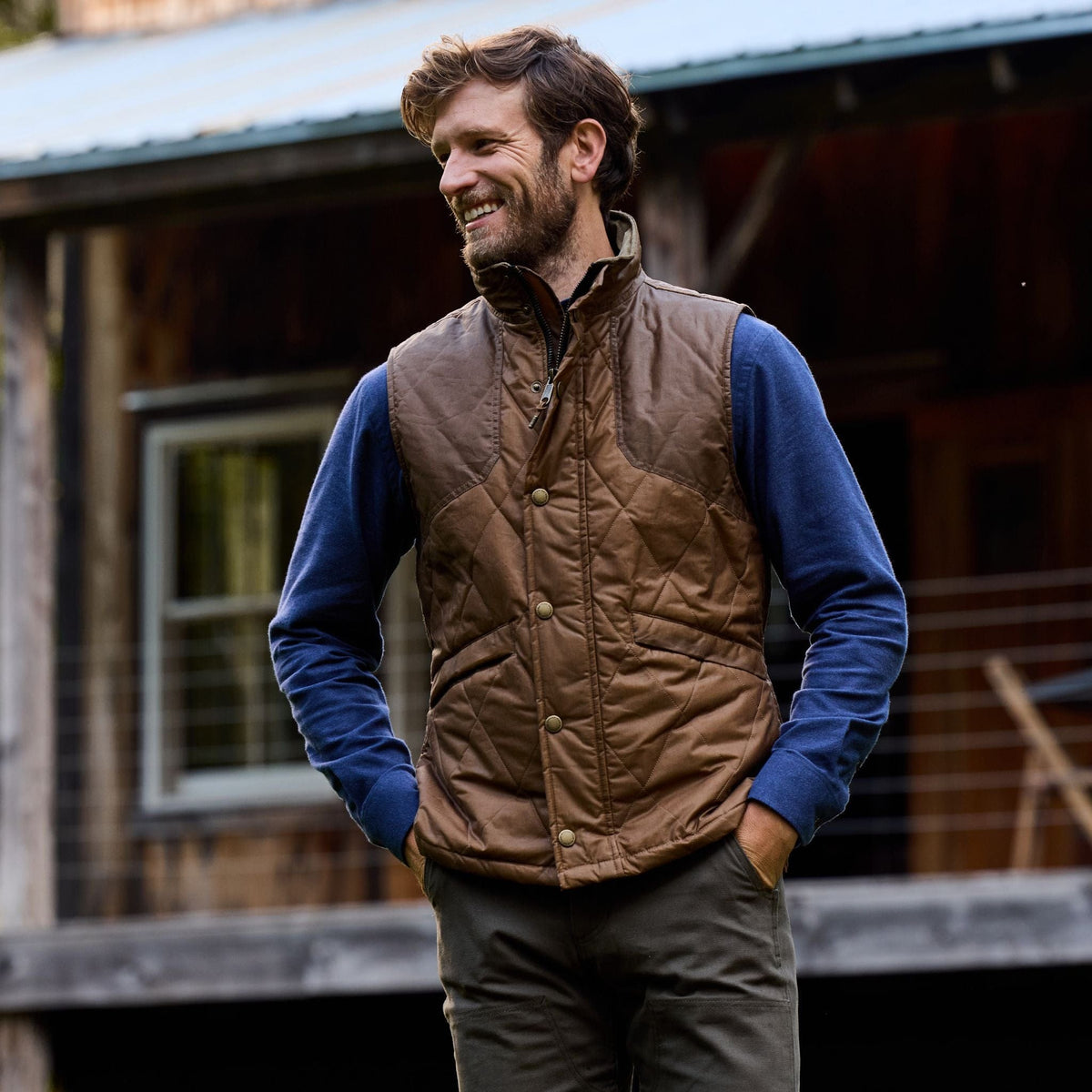 Man in a Tom Beckbe Paddock Quilted Vest and blue shirt stands smiling by a rustic cabin, hands in pockets.