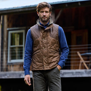 A man stands outside a wooden cabin in a blue shirt and Tom Beckbes Paddock Quilted Vest, made of weatherproof waxed cotton.