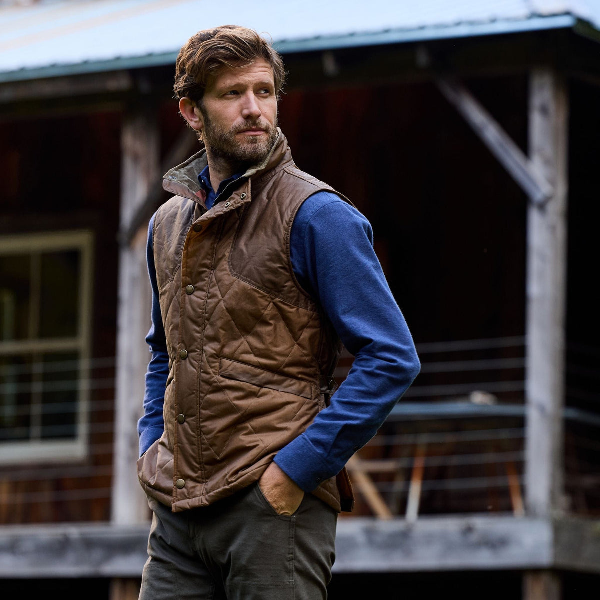 A man with a beard in a Tom Beckbe Paddock Quilted Vest and blue shirt stands outside, hands in pockets, looking sideways.