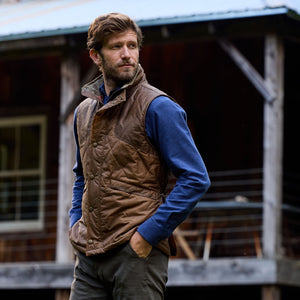 A man with a beard in a Tom Beckbe Paddock Quilted Vest and blue shirt stands outside, hands in pockets, looking sideways.