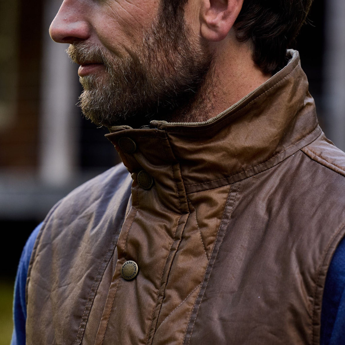 A bearded man in a Tom Beckbe Paddock Quilted Vest and blue sweater stands outdoors; the HD Wool keeps him cozy.
