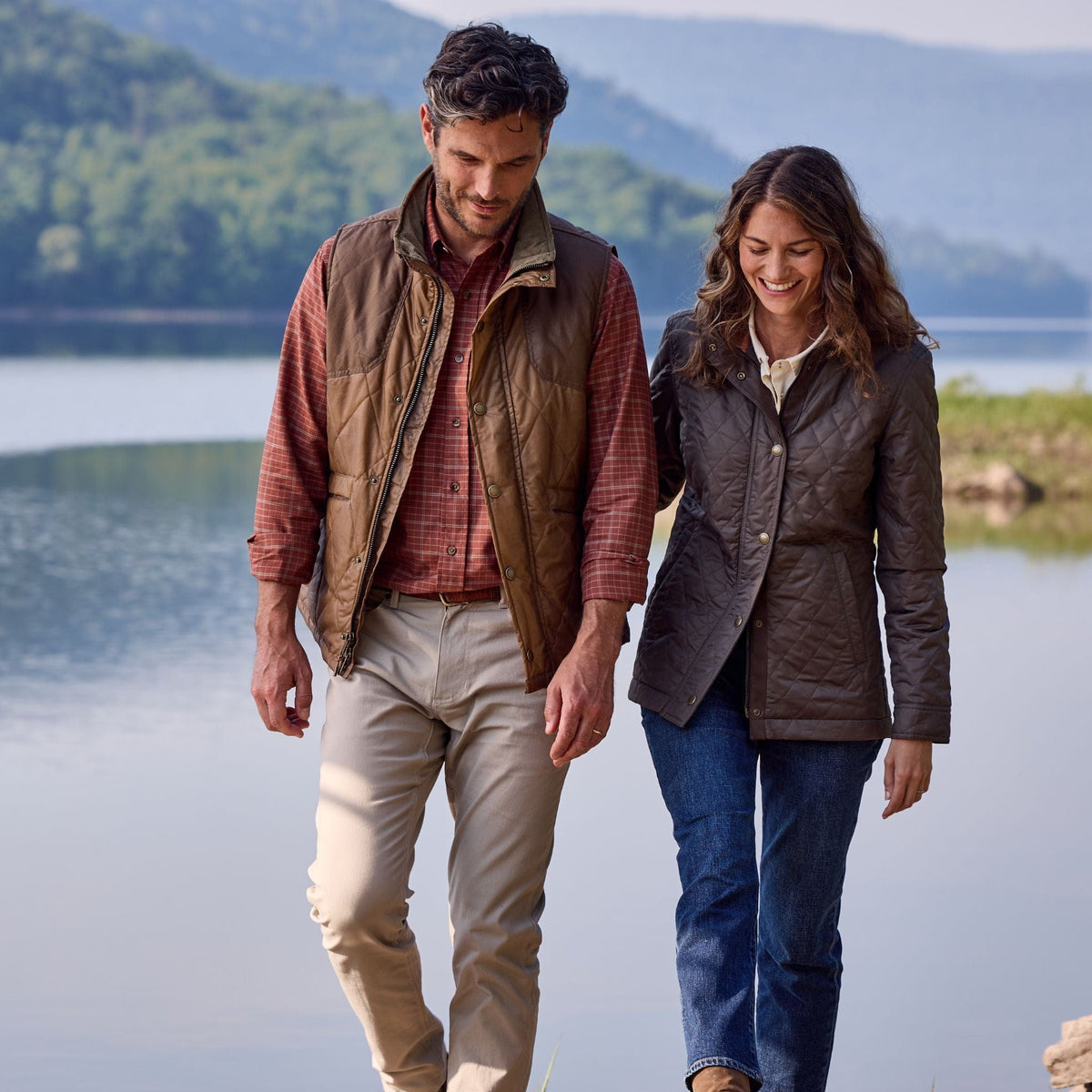 A couple walks lakeside in casual outdoor wear, each wearing a Tom Beckbe Paddock Quilted Vest.