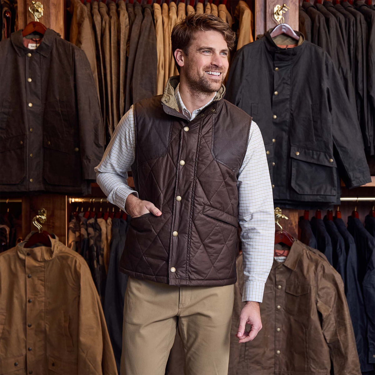 Man wearing a Tom Beckbe Paddock Quilted Vest and khaki pants stands in a store with jackets in the background.
