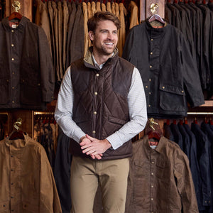 Man wears a Tom Beckbe Paddock Quilted Vest and khaki pants in front of weatherproof vests in a clothing store.